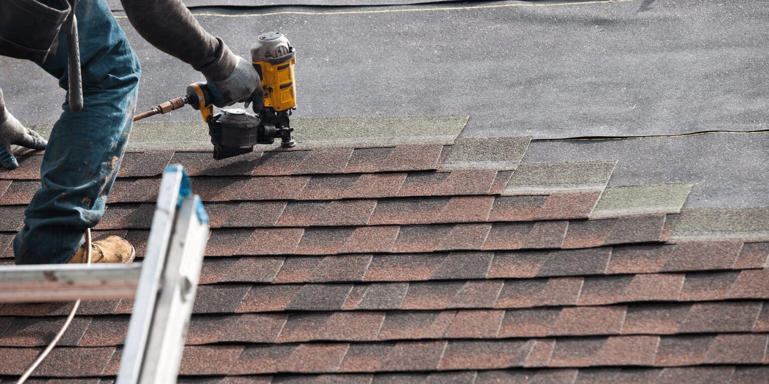 A roofer on a ladder shingling a roof with a nail gun